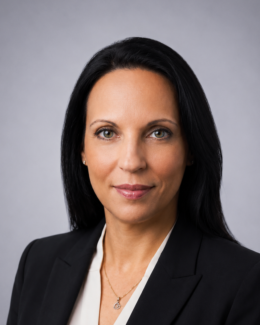 Professional headshot of a woman with dark hair in a black blazer and white blouse, against a gray studio backdrop.