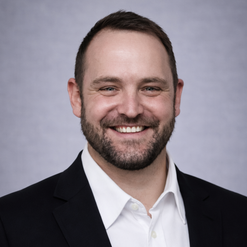 Professional headshot: smiling man in a black blazer and white shirt against a neutral gray backdrop.
