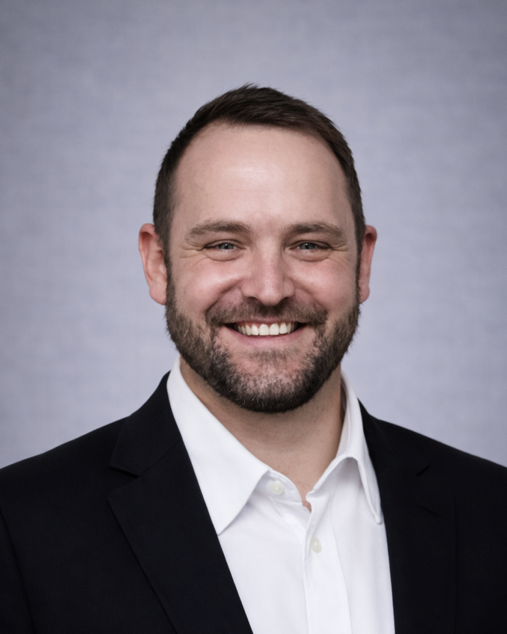 Professional headshot: smiling man in a black blazer and white shirt against a neutral gray backdrop.