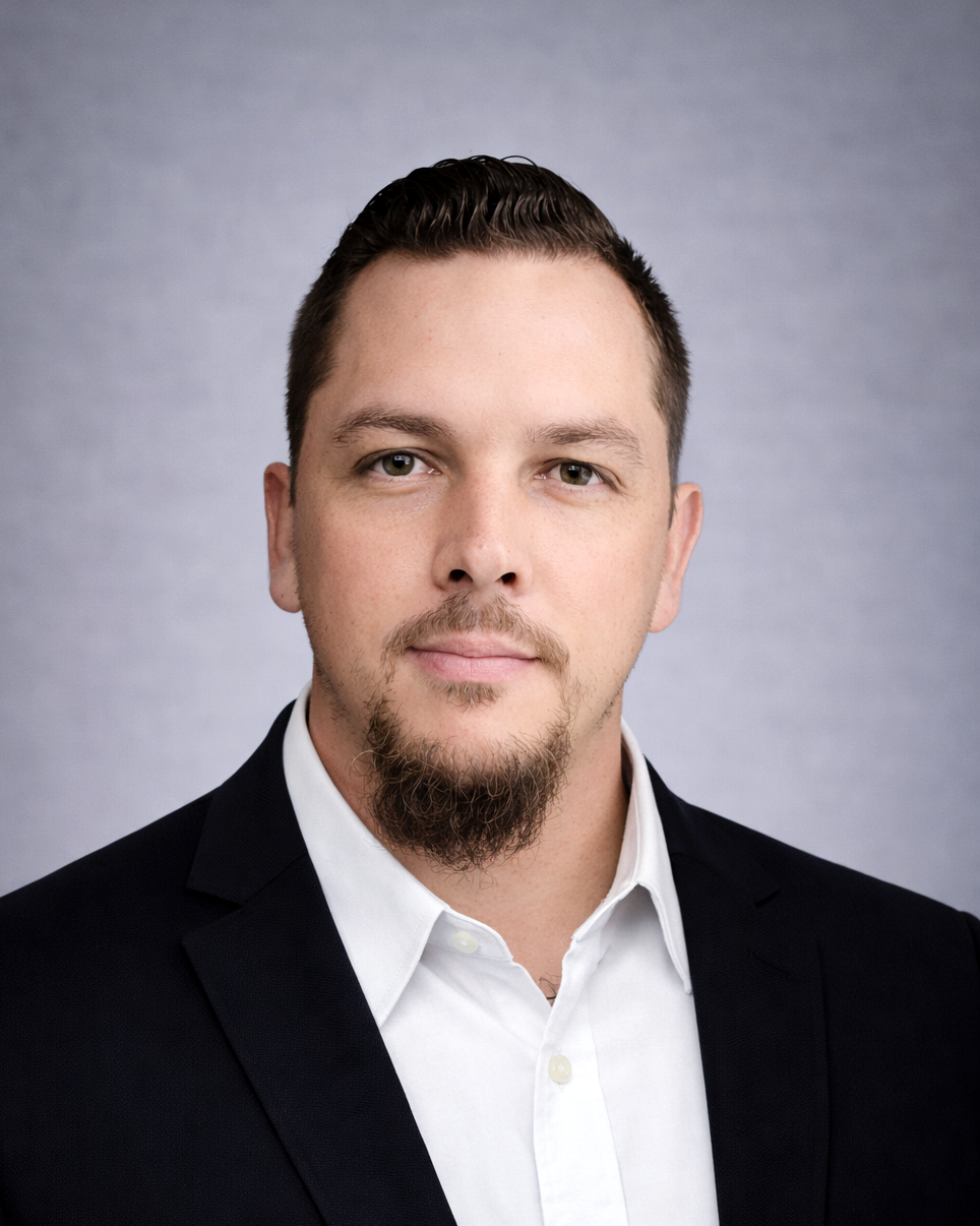 Professional headshot of a man with short dark hair and a goatee wearing a black blazer and white shirt, facing the camera against a gray backdrop.