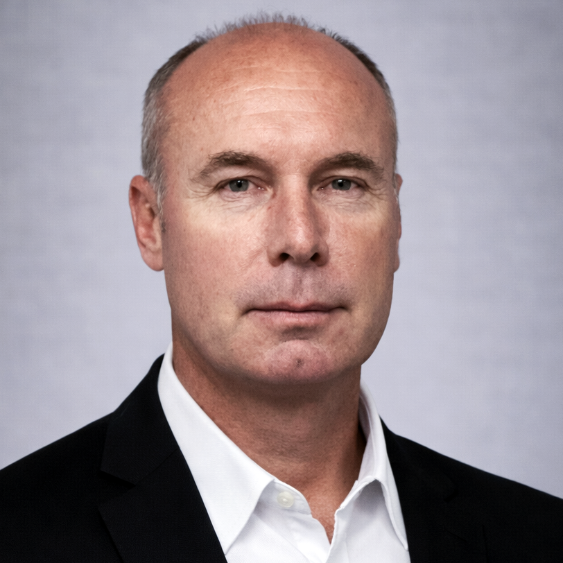 Head-and-shoulders portrait of a bald man in a black suit and white shirt against a light gray background.
