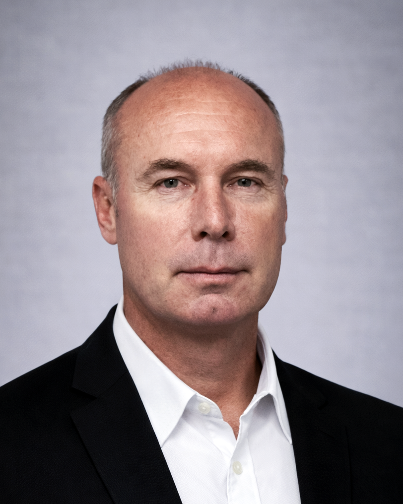 Head-and-shoulders portrait of a bald man in a black suit and white shirt against a light gray background.