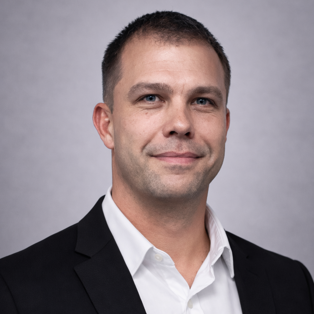 Professional headshot of a smiling man in a black suit jacket and white shirt against a gray backdrop.