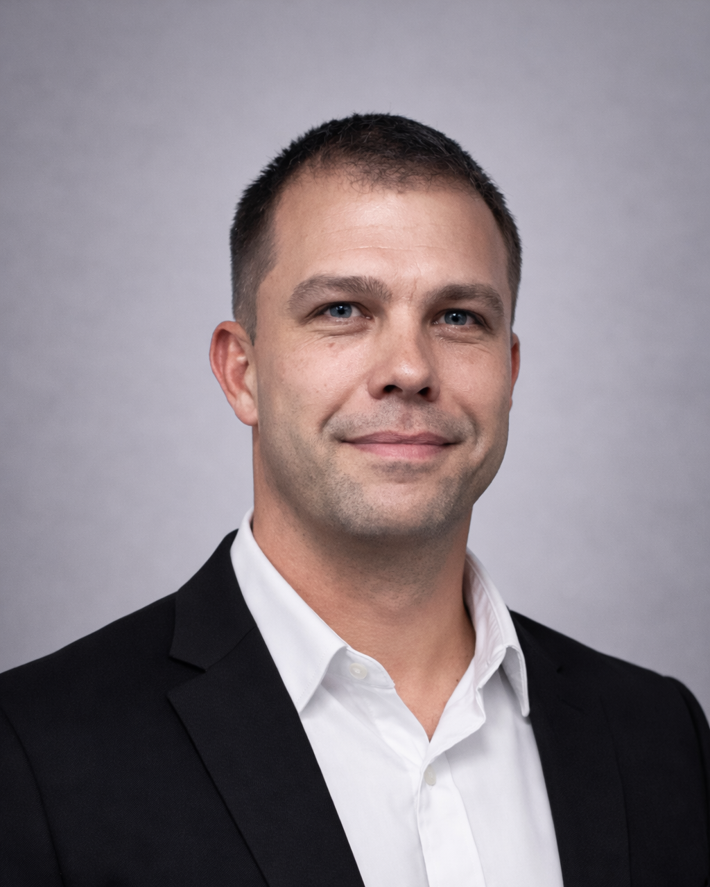 Professional headshot of a smiling man in a black suit jacket and white shirt against a gray backdrop.