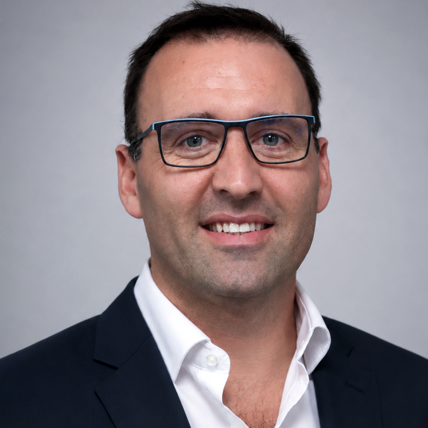 Professional headshot of a man with dark hair and glasses, wearing a navy blazer and white shirt, smiling against a gray backdrop.