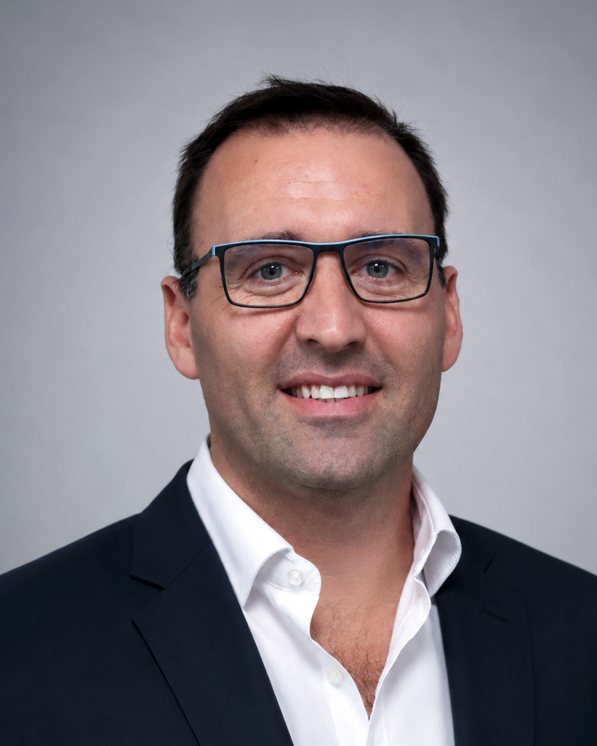 Professional headshot of a man with dark hair and glasses, wearing a navy blazer and white shirt, smiling against a gray backdrop.